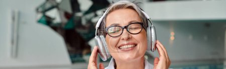 glückliche Frau mittleren Alters mit Brille, die Musik in drahtlosen Kopfhörern im Kurzentrum hört, Banner
