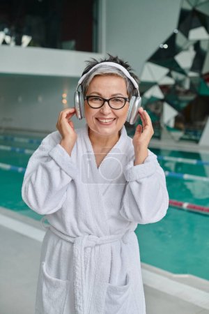 happy middle aged woman in glasses listening music in wireless headphones in spa center, retreat