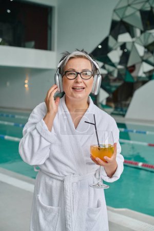 glückliche Frau mittleren Alters hört Musik in drahtlosen Kopfhörern und hält Cocktail im Wellness-Center