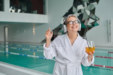 pleased middle aged woman listening music in wireless headphones and holding cocktail in spa center