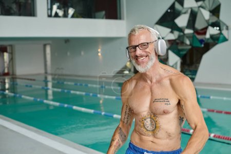 zufriedener und tätowierter Mann mittleren Alters hört Musik in drahtlosen Kopfhörern im Kurzentrum