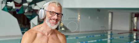 Photo for Pleased and tattooed mature man listening music in wireless headphones in spa center, banner - Royalty Free Image