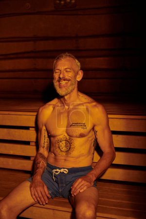 hombre de mediana edad feliz y sin camisa con tatuajes sentado en la sauna, concepto de retiro de bienestar