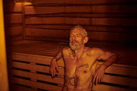Foto de Hombre de mediana edad tranquilo, relajado y sin camisa con tatuajes sentado en la sauna, retiro de bienestar - Imagen libre de derechos