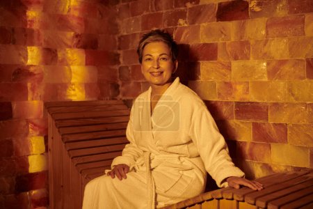 glückliche Frau mittleren Alters im weißen Gewand sitzt auf Bank in Sauna, Wellness-Konzept, Rückzug