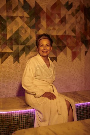 Foto de Sonriente mujer de mediana edad en bata blanca sentada en el banco en la sauna, spa concepto de bienestar, retiro - Imagen libre de derechos