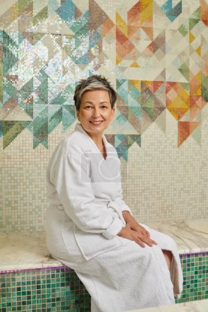 smiling middle aged woman in white robe sitting on bench in marble sauna, spa wellness concept