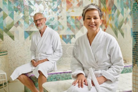 happy middle aged couple in white robes sitting together in marble sauna, spa wellness concept