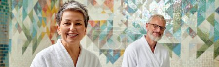 glückliches Paar mittleren Alters in weißen Roben, die zusammen in Marmorsauna, Wellness-Spa, Banner sitzen