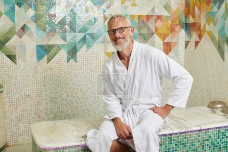 Foto de Alegre hombre de mediana edad en bata blanca sentado en sauna, spa y concepto de bienestar, hammam de mármol - Imagen libre de derechos
