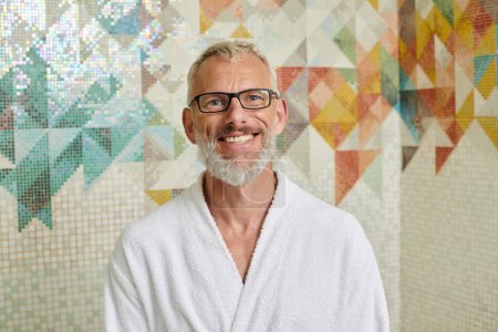Téléchargez les photos : Joyeux homme d'âge moyen en robe blanche assis dans le sauna, concept spa et bien-être, hammam en marbre - en image libre de droit