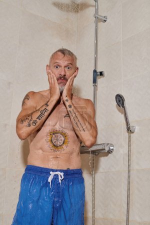 Foto de Divertido hombre de mediana edad con tatuajes tomando ducha y lavando la cara, higiene personal - Imagen libre de derechos