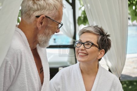 glückliche Frau mittleren Alters in Brille schaut Ehemann in weißer Robe an, Wellness-Rückzugskonzept