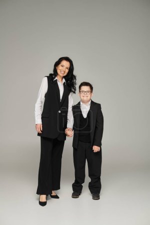 Téléchargez les photos : Femme heureuse en tenue formelle tenant la main de son fils avec le syndrome du duvet en uniforme scolaire sur gris - en image libre de droit