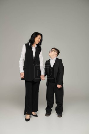 Geschäftsfrau und Sohn mit Down-Syndrom halten Händchen, lächeln einander auf grauer, voller Länge an