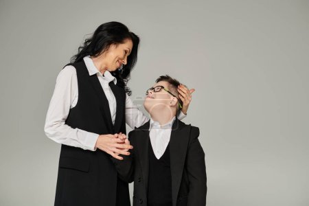 Téléchargez les photos : Femme en tenue de travail et fils avec le syndrome du duvet en uniforme scolaire se regardant sur gris - en image libre de droit