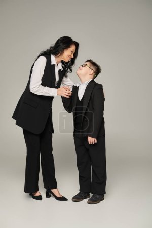 Téléchargez les photos : Femme d'affaires et fils avec le syndrome du duvet en uniforme scolaire tenant la main et souriant sur gris - en image libre de droit