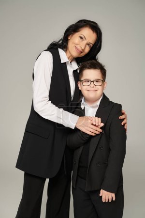 woman in formal wear and happy boy with down syndrome in school uniform looking at camera on grey