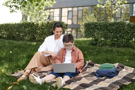 Téléchargez les photos : Garçon avec le syndrome du duvet, dans les lunettes, assis avec ordinateur portable dans le parc près de mère heureuse, e-learning - en image libre de droit