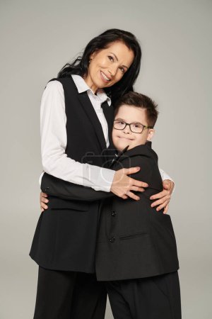 Photo for Cheerful schoolboy with down syndrome embracing mother in formal wear and looking at camera on grey - Royalty Free Image