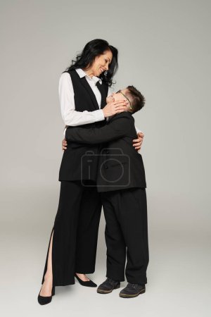 boy with down syndrome, in school uniform, and woman in formal wear embracing on grey, unique family
