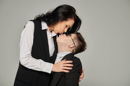 woman in formal wear kissing son with down syndrome in school uniform on grey, unconditional love