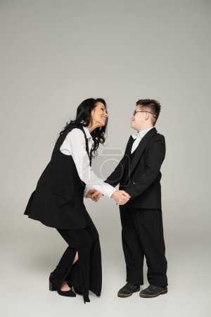 joyful businesswoman holding hands of son with down syndrome in school uniform on grey, full length