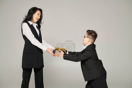 kid with down syndrome giving present to amazed mother in elegant outfit on grey, unique family