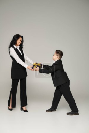 Junge mit Down-Syndrom in Schuluniform überreicht Geschenk an glückliche Mutter in formalem Outfit auf grau