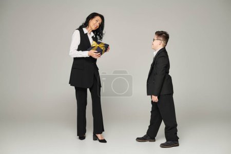 Frau in Geschäftskleidung hält Geschenkbox neben Sohn mit Down-Syndrom in Schuluniform auf grau