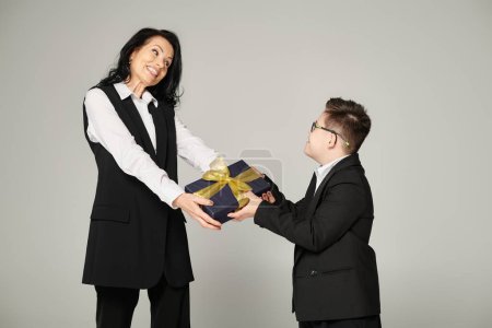 freudige Geschäftsfrau überreicht Geschenk an Sohn mit Down-Syndrom in Schuluniform auf grau