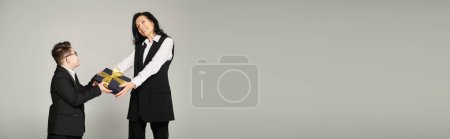 Téléchargez les photos : Femme en tenue formelle donnant boîte cadeau à son fils avec le syndrome du duvet en uniforme scolaire sur gris, bannière - en image libre de droit