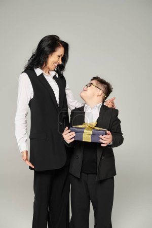 pleased schoolboy with down syndrome holding present near mother in formal wear on grey, happiness