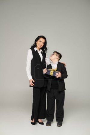 Frau in Business-Kleidung lächelt in die Kamera neben Sohn mit Down-Syndrom hält Geschenkbox auf grau