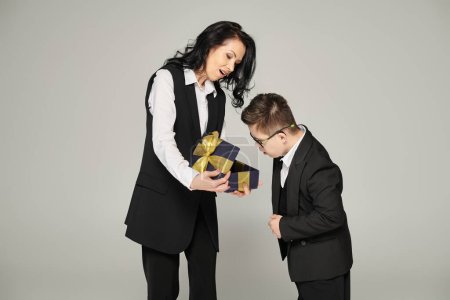 Frau in offizieller Kleidung überreicht Geschenk an überraschten Sohn mit Down-Syndrom in Schuluniform auf grau