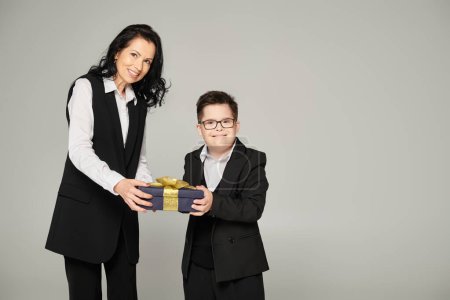 freudige Mutter und Sohn mit Down-Syndrom in Schuluniform mit Geschenkverpackung und grinsendem Lächeln auf grau
