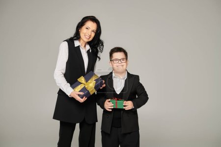 Frau in Businesskleidung und Junge mit Down-Syndrom in Schuluniform mit Geschenkschachteln auf grau