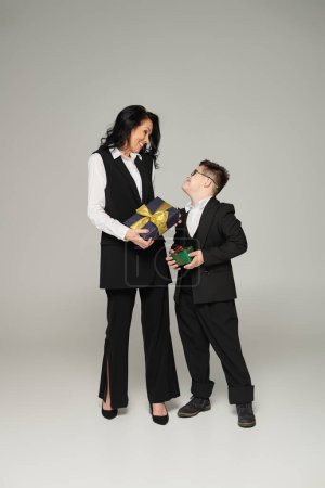 Téléchargez les photos : Femme d'affaires et fils heureux avec le syndrome du duvet en uniforme scolaire tenant des boîtes-cadeaux sur gris - en image libre de droit