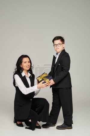 freudige, elegante Mutter und ihr Sohn mit Down-Syndrom halten eine Geschenkschachtel in der Hand und blicken in die Kamera auf grau