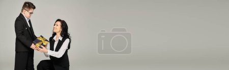 Téléchargez les photos : Femme souriante en tenue de cérémonie présentant un cadeau au fils atteint du syndrome du duvet sur gris, bannière - en image libre de droit