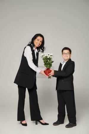 glückliche Geschäftsfrau und Sohn mit Down-Syndrom mit Geschenkschachtel und Blumen auf grau