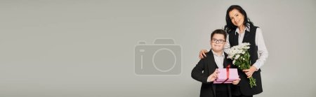 Téléchargez les photos : Femme d'affaires souriante et fils avec le syndrome du duvet tenant des fleurs et boîte cadeau sur gris, bannière - en image libre de droit