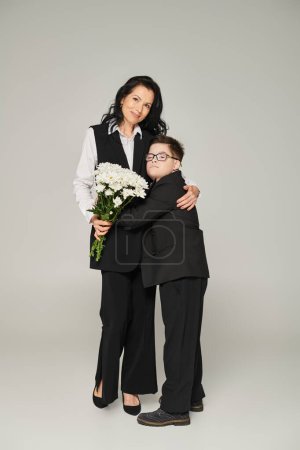 glückliche Frau und Junge mit Down-Syndrom, die Blumen in der Hand halten und sich auf einer grauen, einzigartigen Familie umarmen