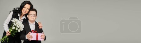 Téléchargez les photos : Mère et garçon souriants avec syndrome duvet tenant boîte cadeau et fleurs sur gris, bannière - en image libre de droit