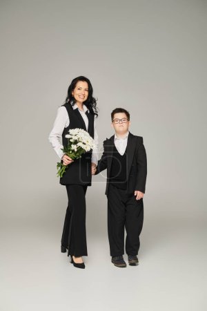 Téléchargez les photos : Femme en tenue formelle tenant des fleurs près de son fils avec le syndrome du duvet en uniforme scolaire sur gris - en image libre de droit