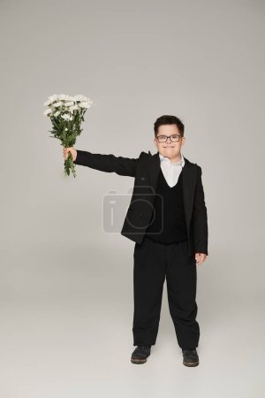 schoolboy with down syndrome in eyeglasses and uniform holding bouquet in outstretched hand on grey
