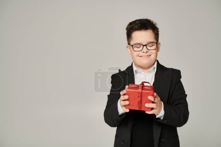 pleased schoolboy with mental disability holding red gift box on grey, down syndrome concept