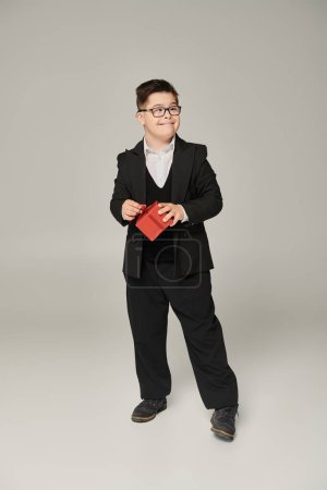 fröhlicher Junge mit Down-Syndrom in Schuluniform und Brille mit rotem Geschenkkarton auf grau