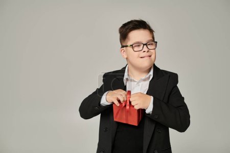 Kind mit Down-Syndrom in Brille und Schuluniform mit Geschenkverpackung auf grau, Sonderschüler
