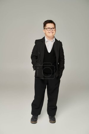 schoolboy with mental disability posing in black suit with hands in pockets on grey